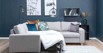 Lys grå sovesofa foran mørk blå væg med dekorative puder, vægkunst og glaspendel.