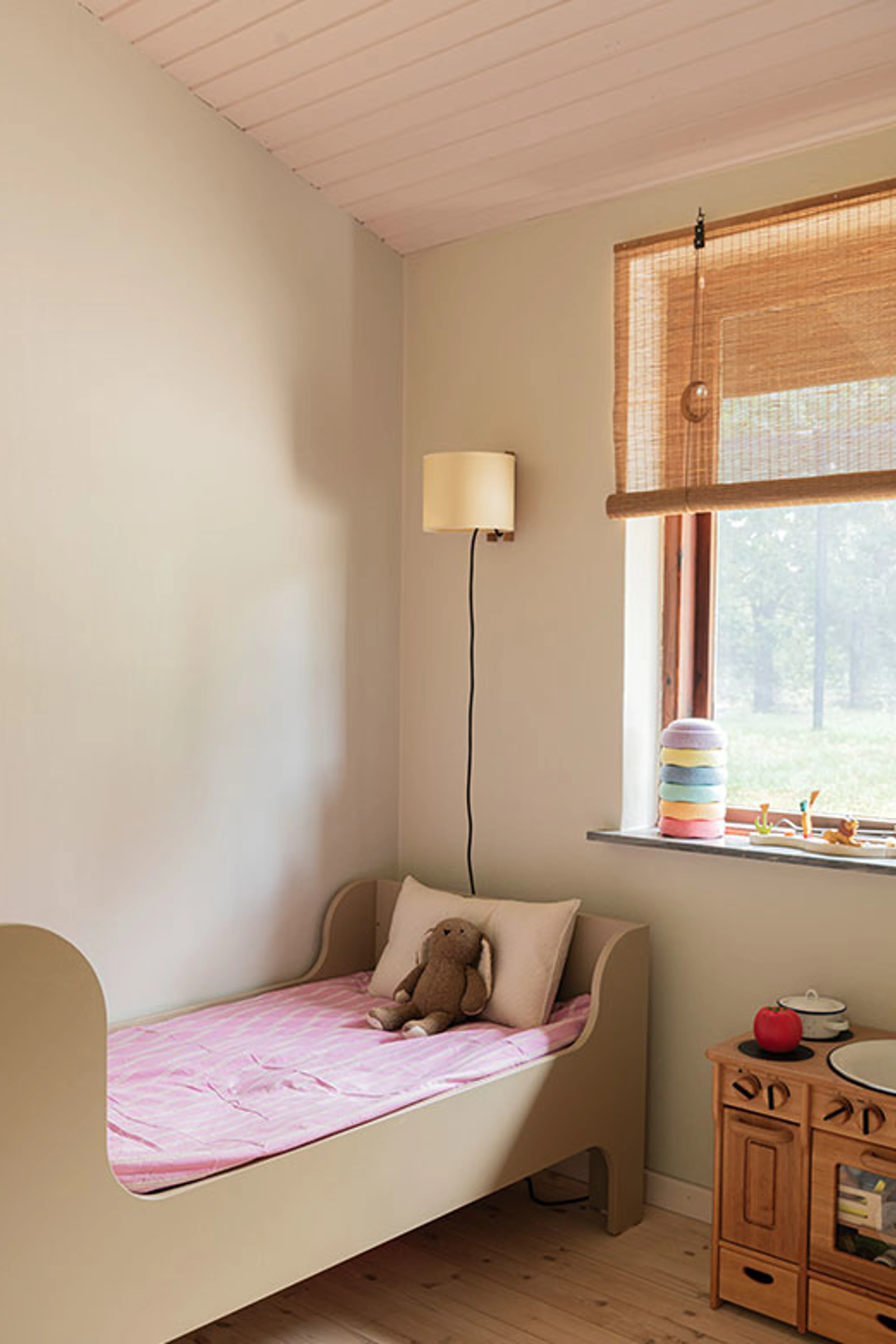 Children's room with beige walls, a small bed with pink bedding and a bamboo blind.