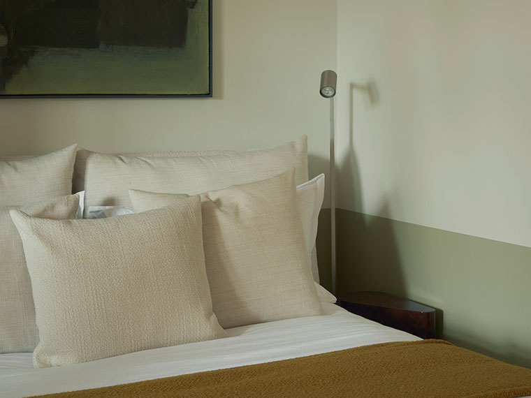 Detail of a made bed with cream-colored pillows, a mustard yellow throw, and a wall in muted shades of green.