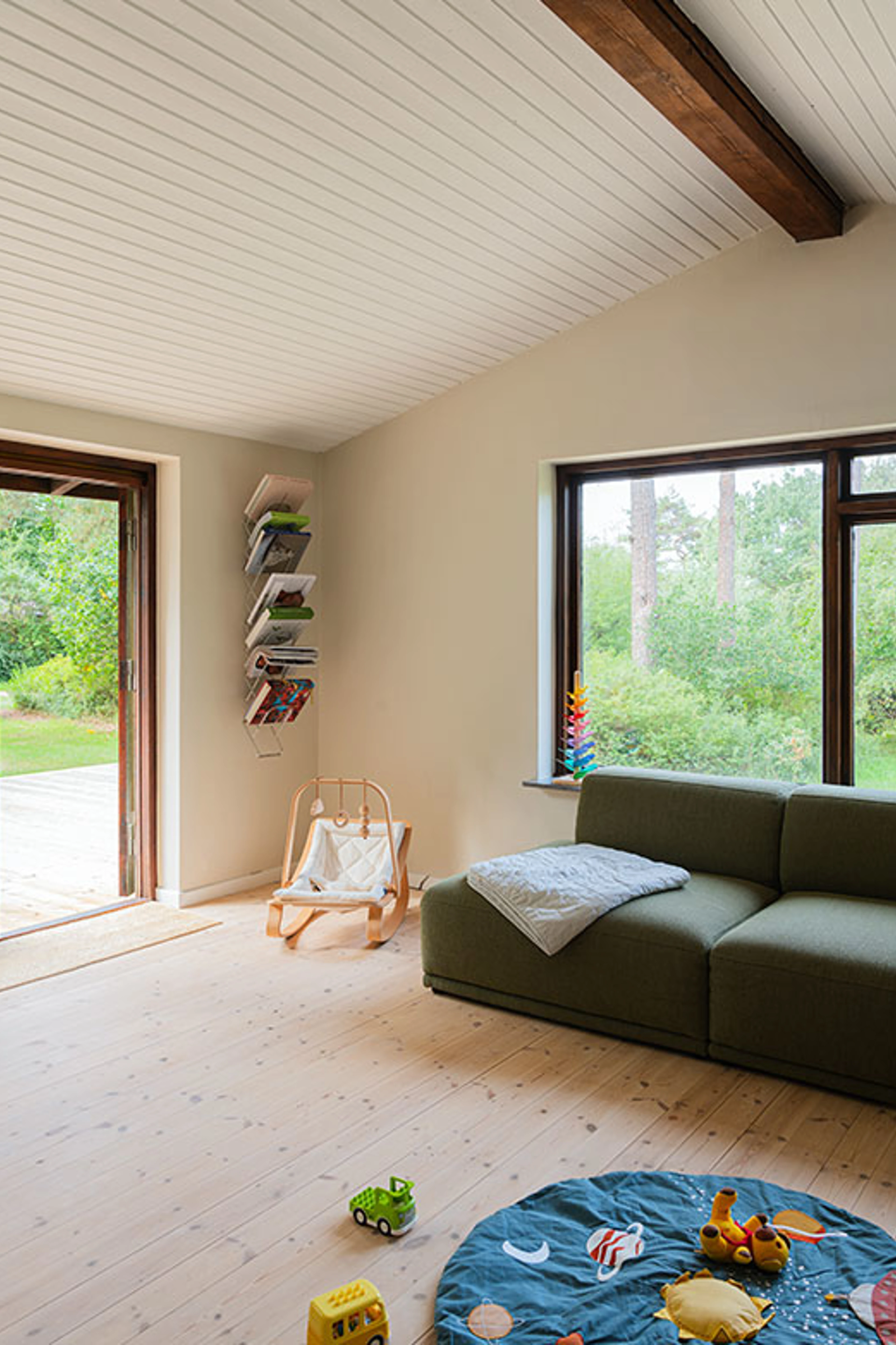Bright living room with beige walls, a white ceiling with a dark beam, a green sofa and a large window facing the garden.