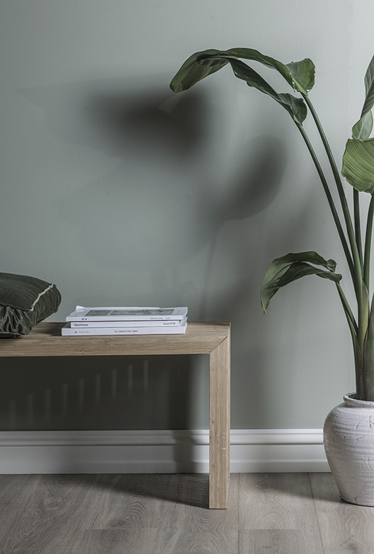 Lys grågrøn væg med træbænk, bøger og stor grøn potteplante. Rummet er indrettet i naturlige materialer og afdæmpede farver.