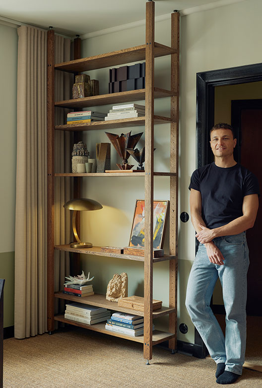 Man stands beside a tall dark wood shelving unit with books, art objects, and a brass lamp.