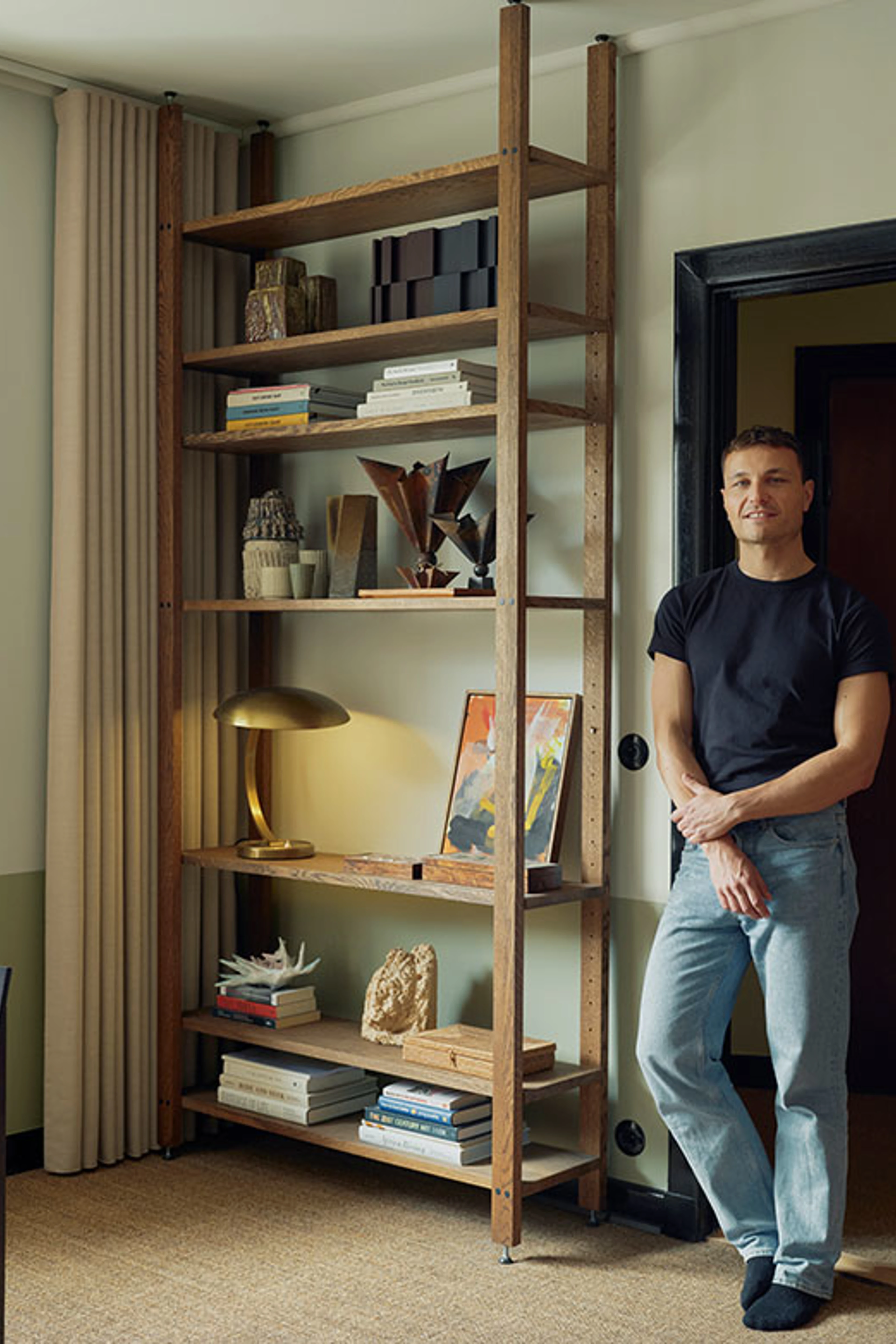 Man stands beside a tall dark wood shelving unit with books, art objects, and a brass lamp.