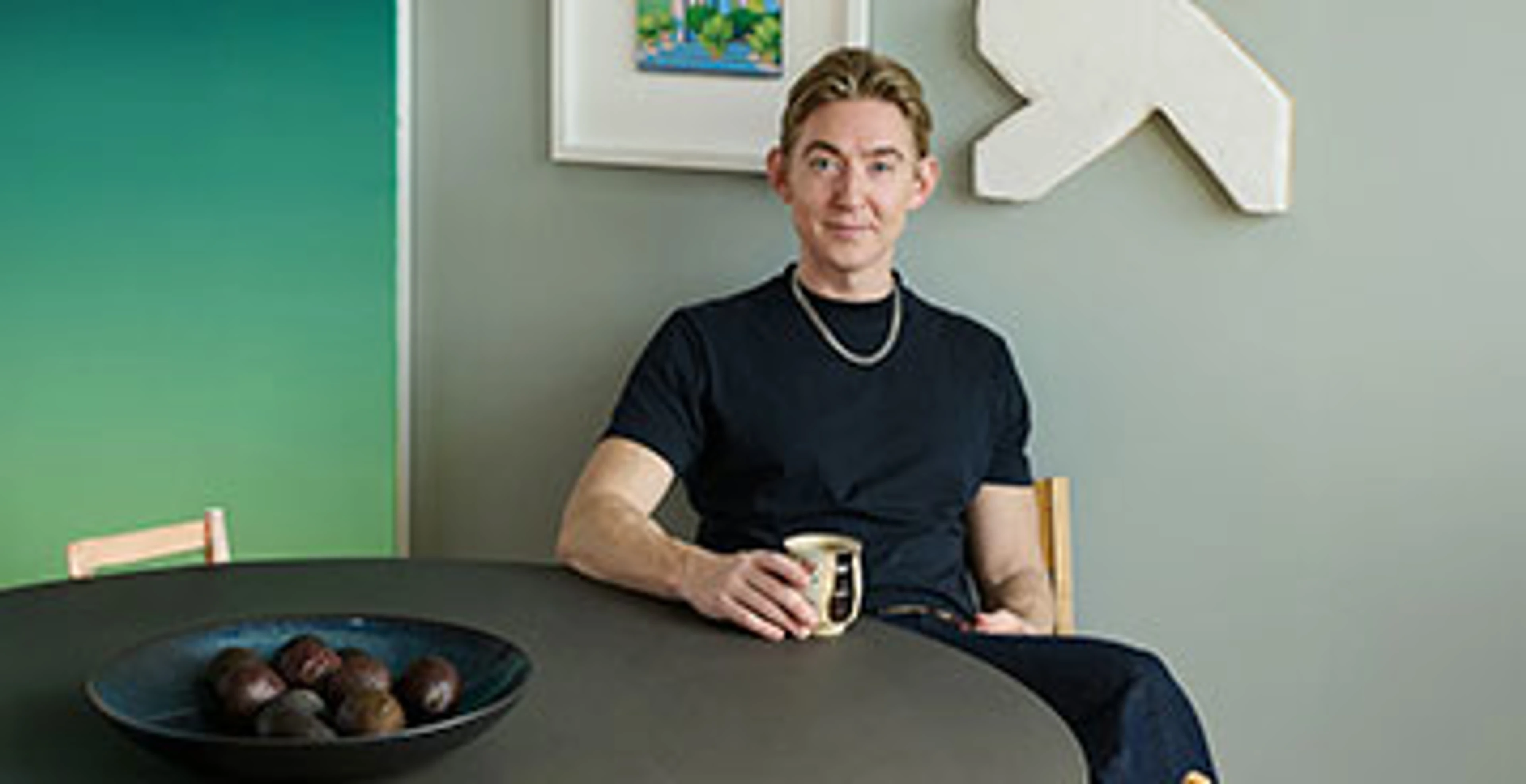Emil Thorups sidder ved mørkt spisebord med en kop i hånden og en grøn væg bag sig.