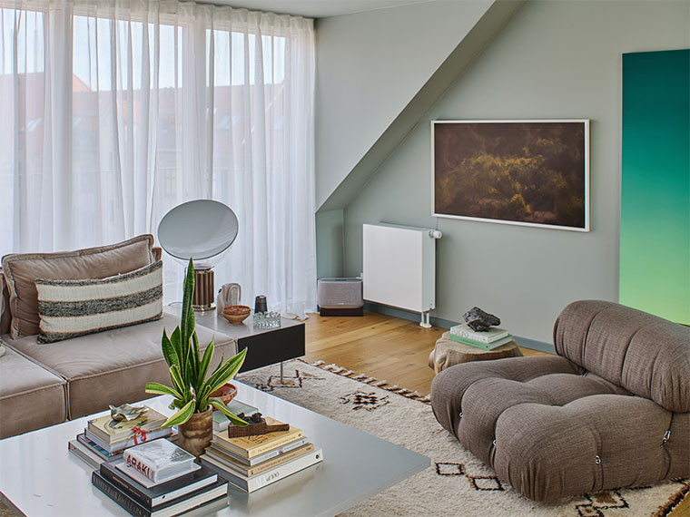 Stue med lyse vægge, sofaer, tæppe og kunst, indrettet med planter og tekstiler i rolige nuancer.
