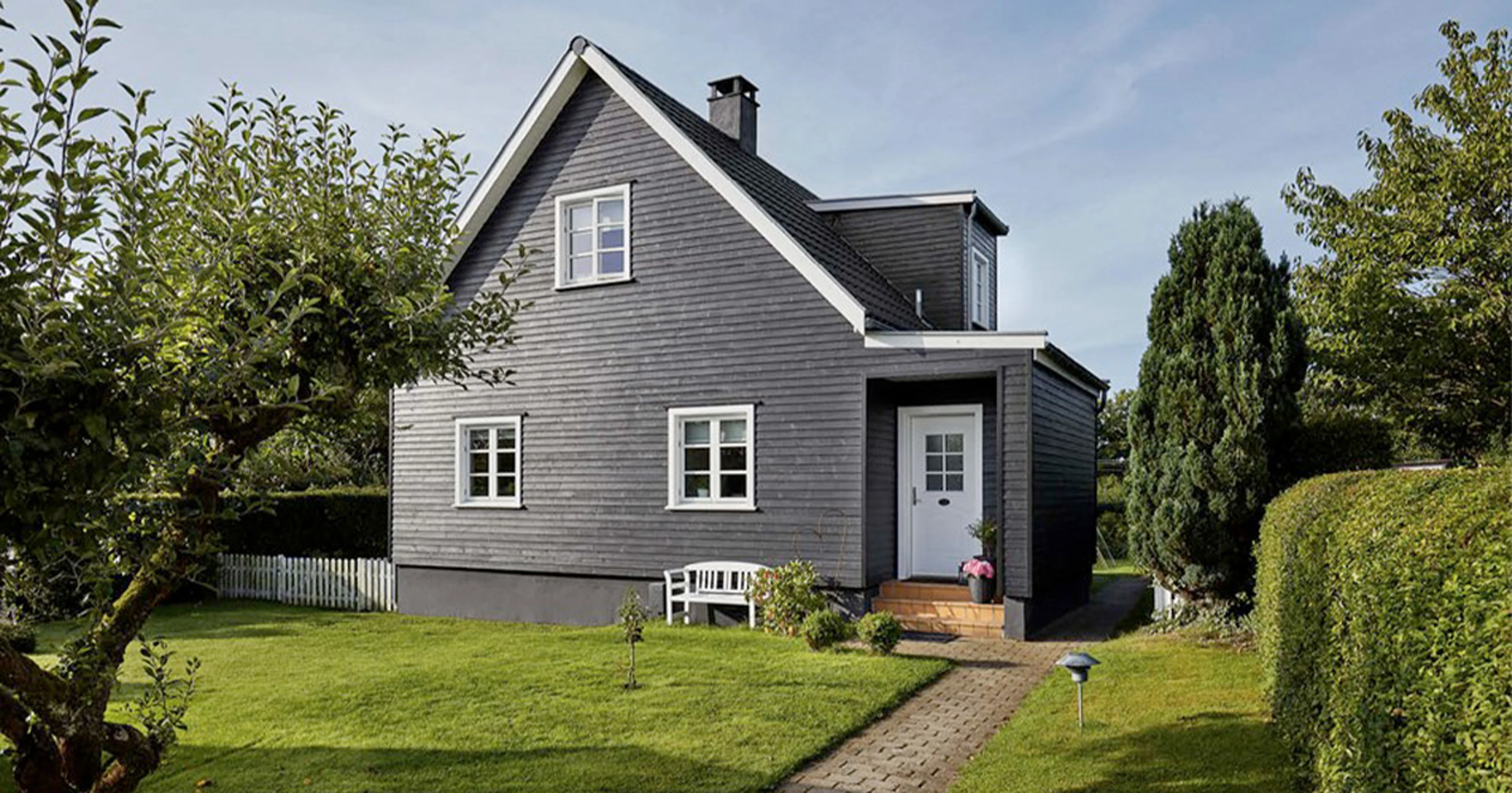 Villa med mørk facade og hvide vinduesrammer omgivet af en velholdt have. En sti fører gennem græsplænen frem til husets indgang.