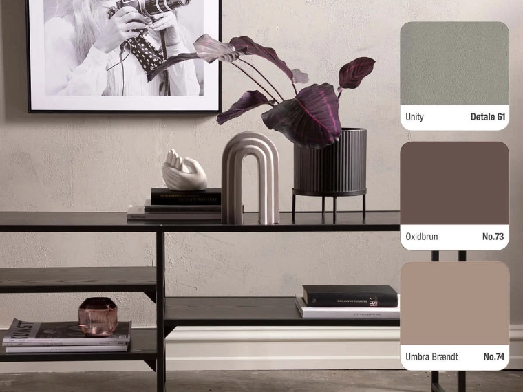 Warm grey wall with a black shelving unit holding a purple-leaved plant in a dark planter, an arch-shaped sculpture, a hand figurine, books and a pink glass candle holder, with a framed black-and-white photograph above and three colour swatches on the right: Unity Detale 61, Oxidbrun No.73 and Umbra Brændt No.74.