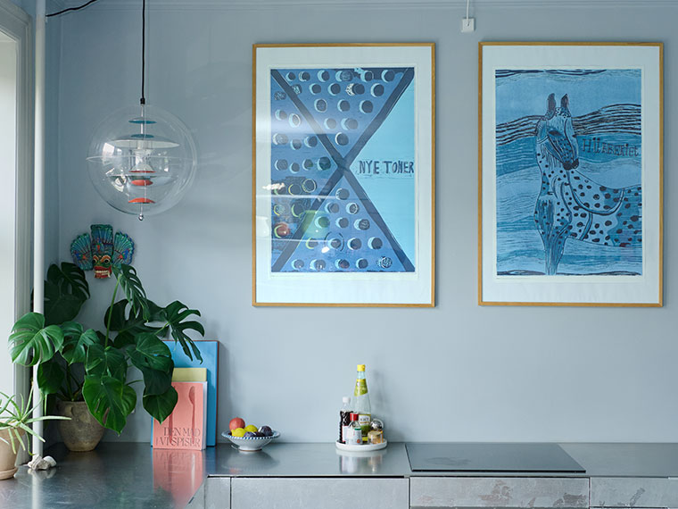 Køkken med stålbordplade, blå kunstplakater, pendellampe i glas og grøn stueplante.
