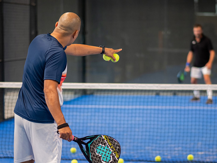 Person står på en indendørs padelbane med et padelbat i hånden og flere gule bolde spredt på banen. En anden spiller ses på den modsatte side af nettet. Billedet viser træning eller instruktion i padel.
