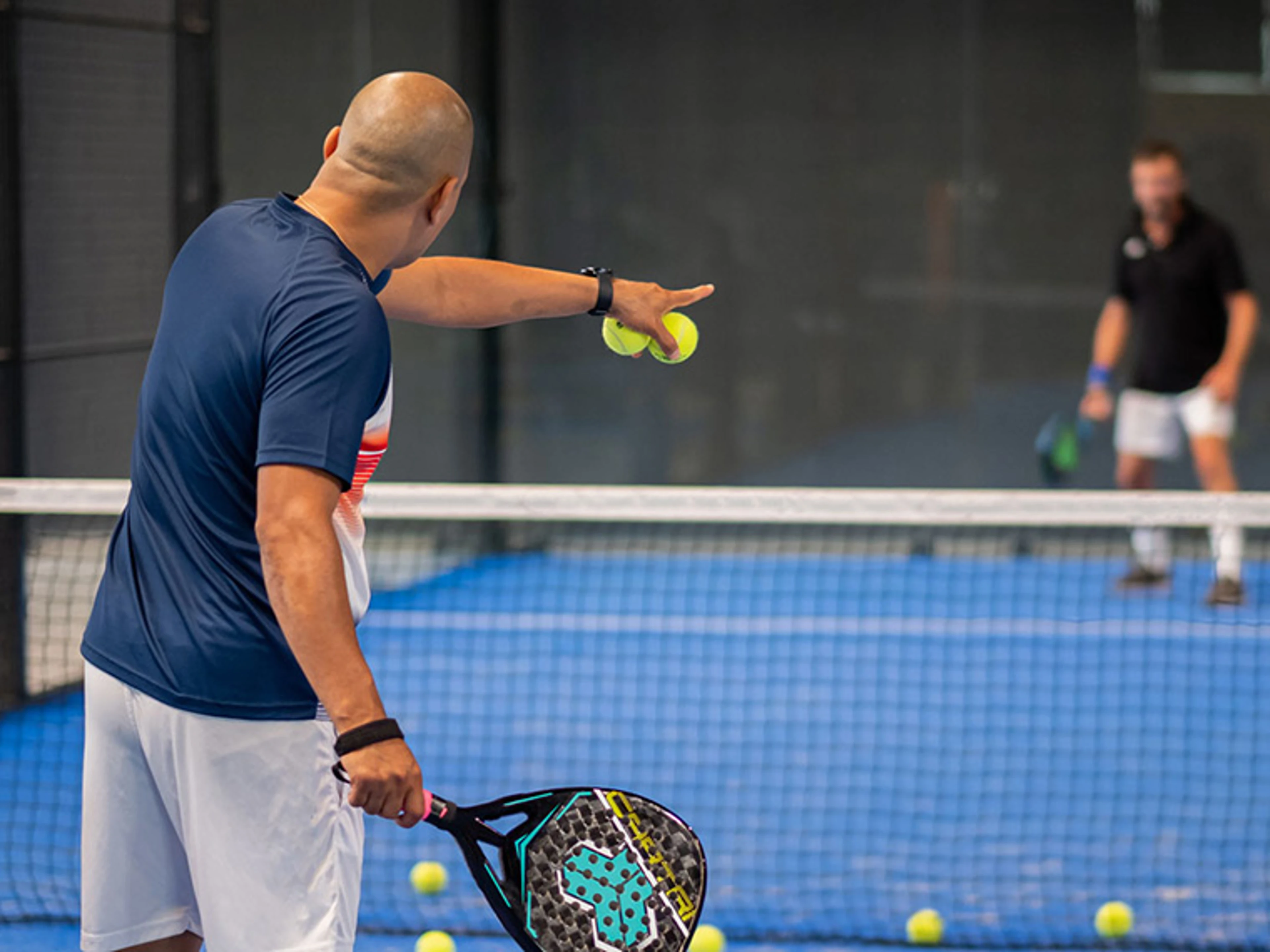 Person står på en indendørs padelbane med et padelbat i hånden og flere gule bolde spredt på banen. En anden spiller ses på den modsatte side af nettet. Billedet viser træning eller instruktion i padel.