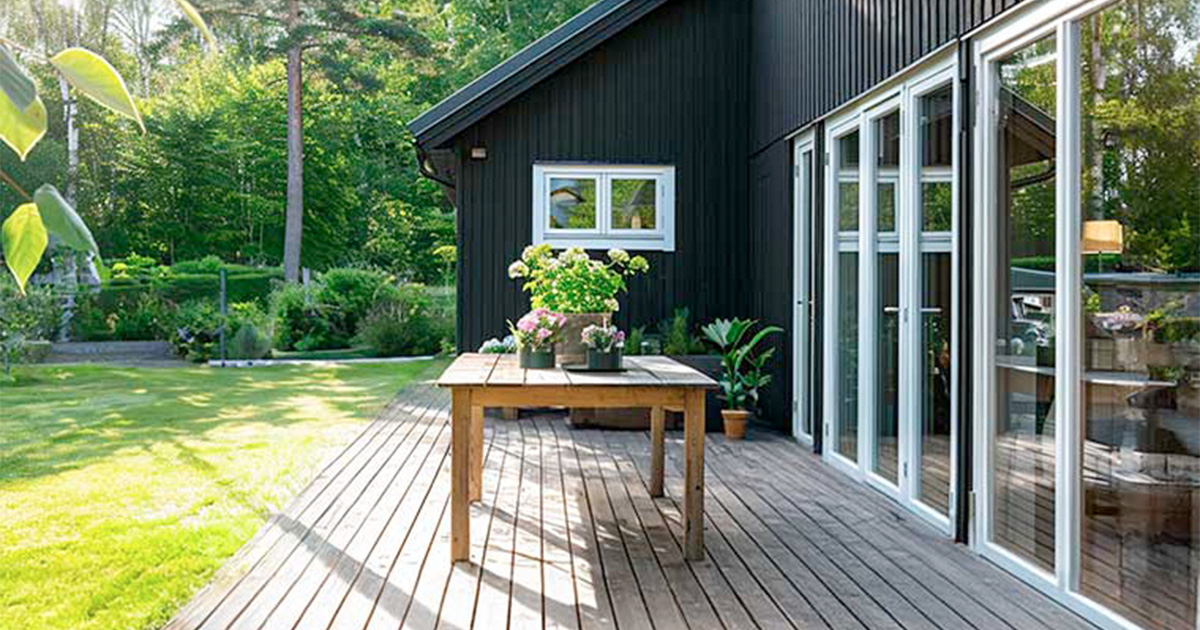Sortmalet træhus med stor træterrasse og glaspartier ud mod en grøn have. På terrassen står et træbord med blomster.