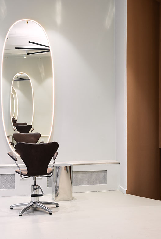 Frisørstol foran stort, ovalt spejl med integreret belysning i minimalistisk salon.