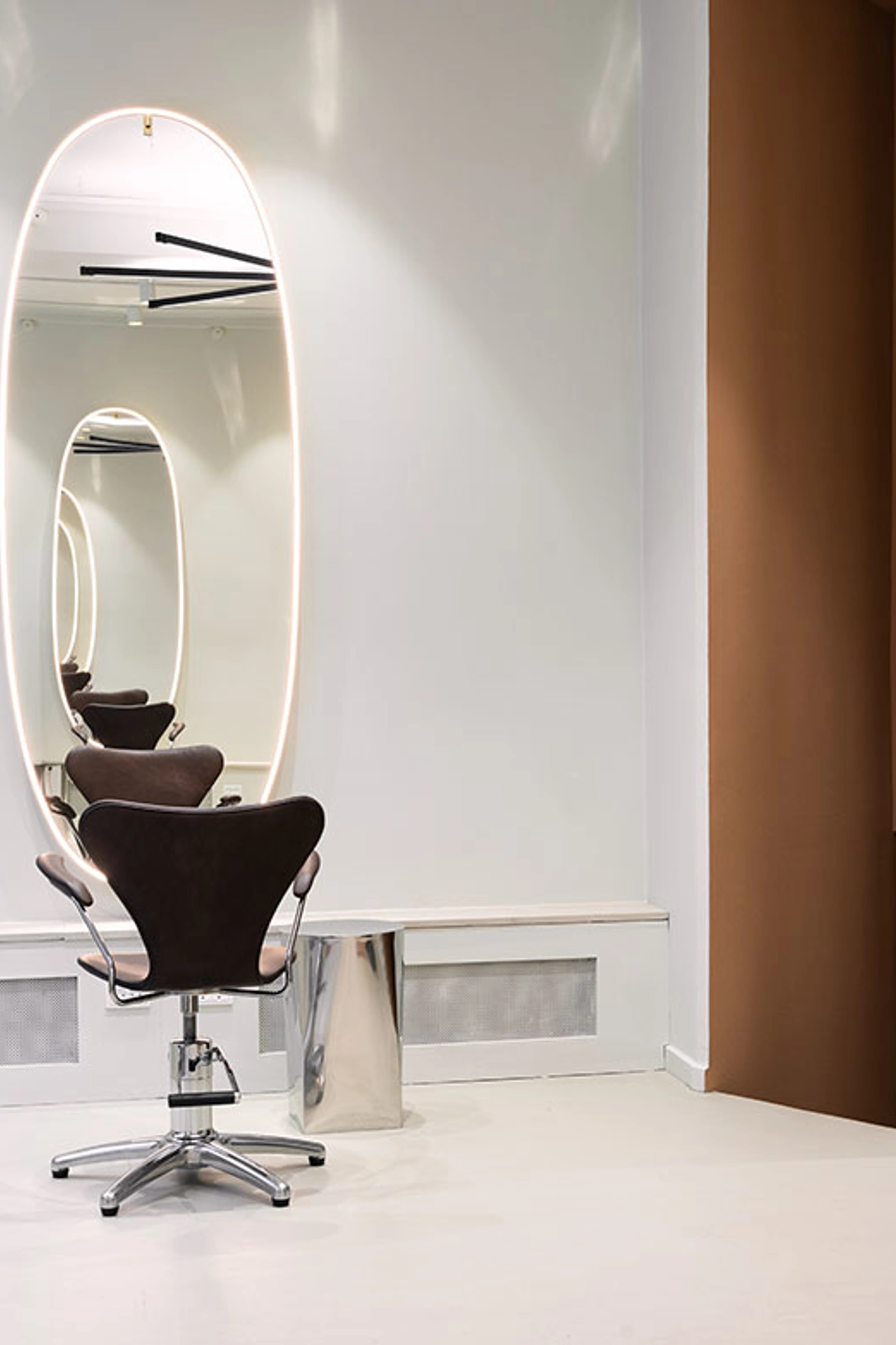 Frisørstol foran stort, ovalt spejl med integreret belysning i minimalistisk salon.