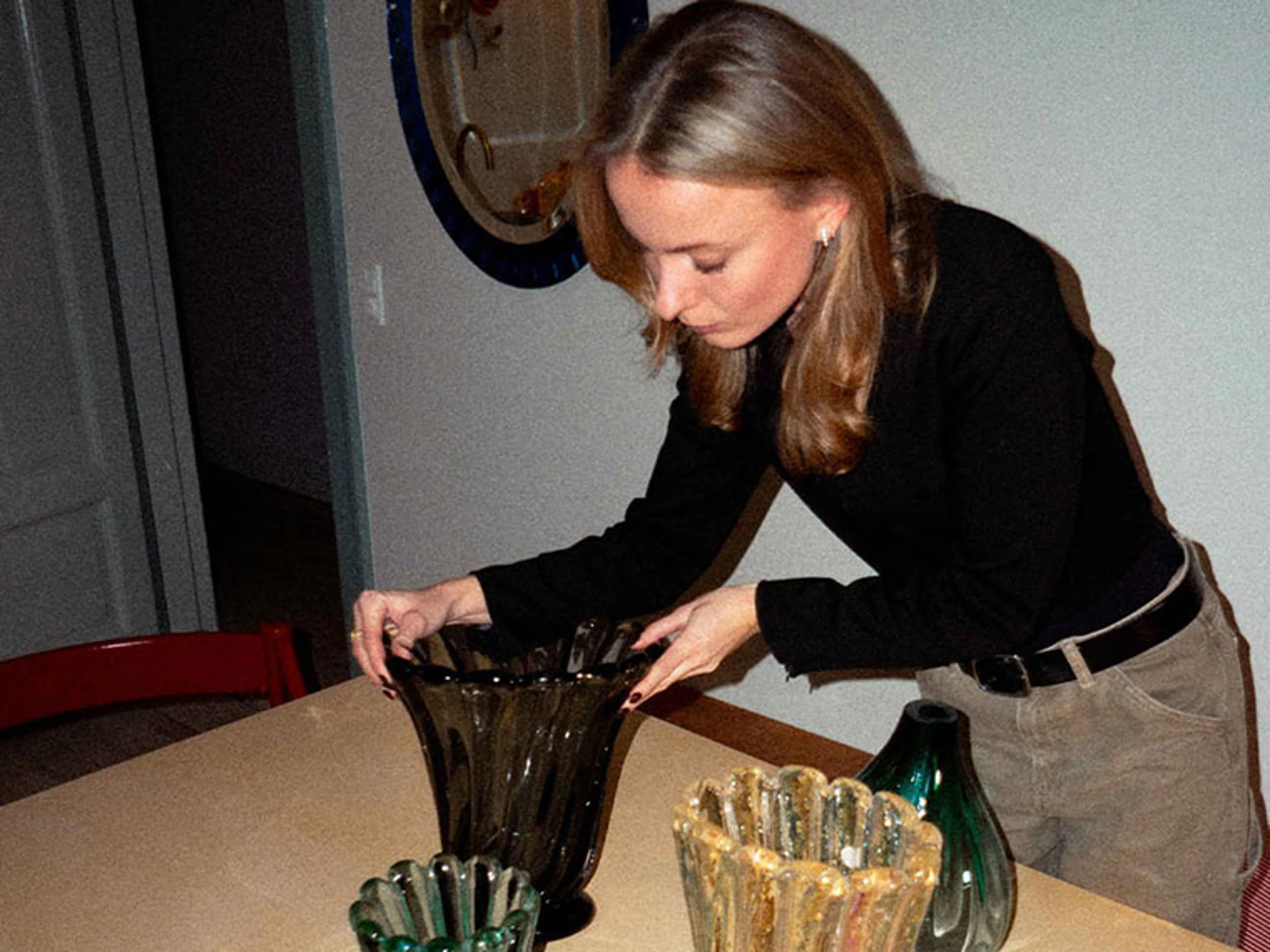 Frederikke arrangerer glasvaser i forskellige farver på et træbord.