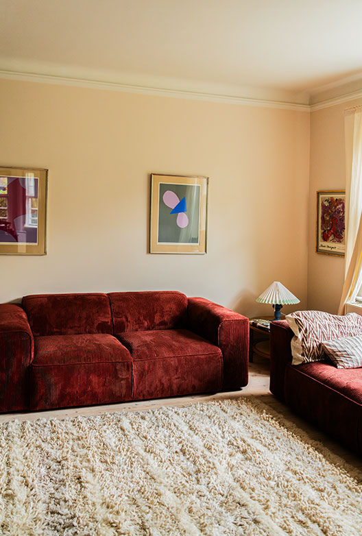 Stue med bordeaux sofa, lyst tæppe og kunst på væggene.