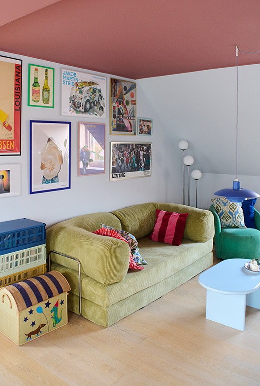 Stue med lyse vægge og rosa loft, indrettet med sofa, farverige puder, sofabord og væg med plakater.