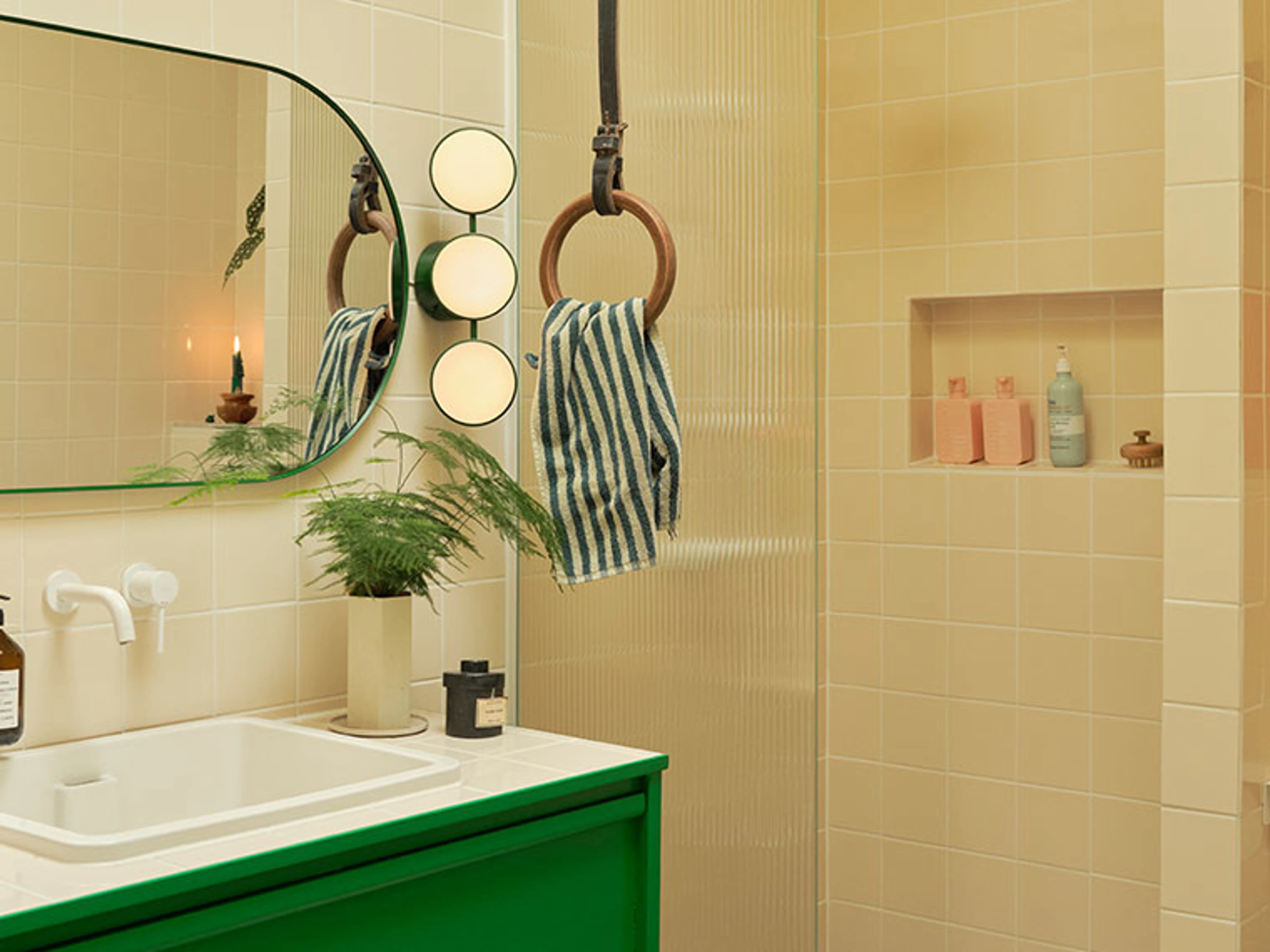 Bathroom with light yellow tiles, a green sink, an organically shaped mirror, round wall lights, and striped towels.