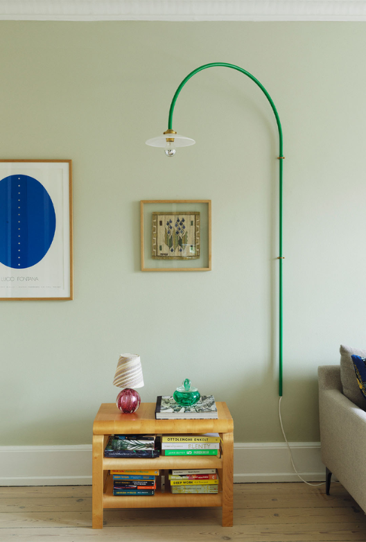 Lysegrøn væg med to malerier samt en væghængt grøn lampe og et firkantet træbord med bøger og en lille lampe. 