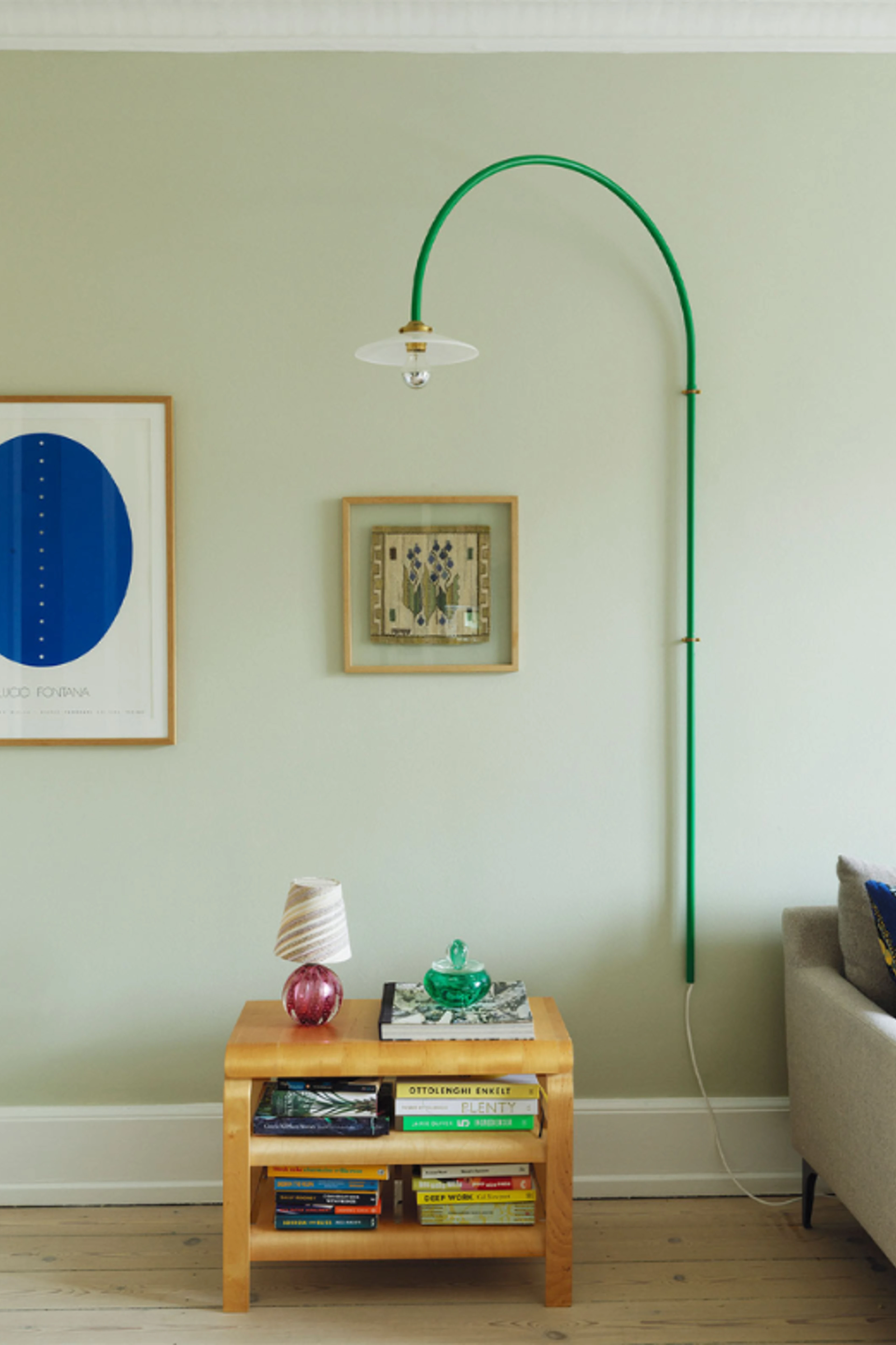 Lysegrøn væg med to malerier samt en væghængt grøn lampe og et firkantet træbord med bøger og en lille lampe.