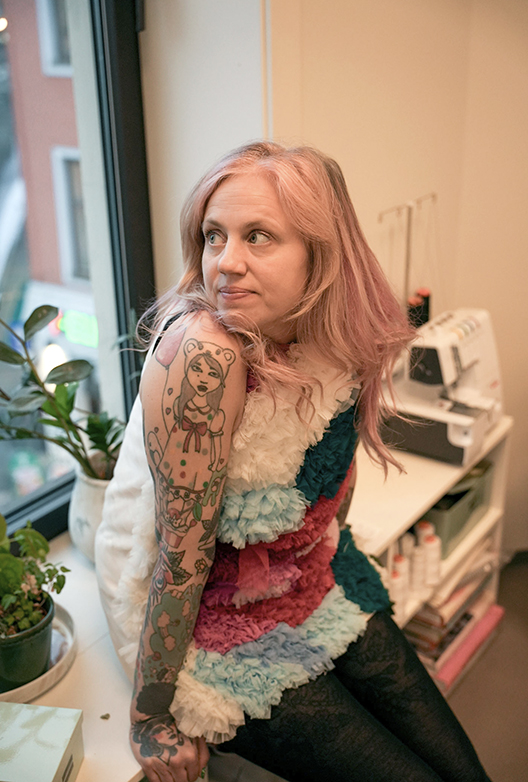 Kvinde med lyserødt hår og tatoverede arme sidder ved et vindue i et arbejdsrum med symaskine og planter.