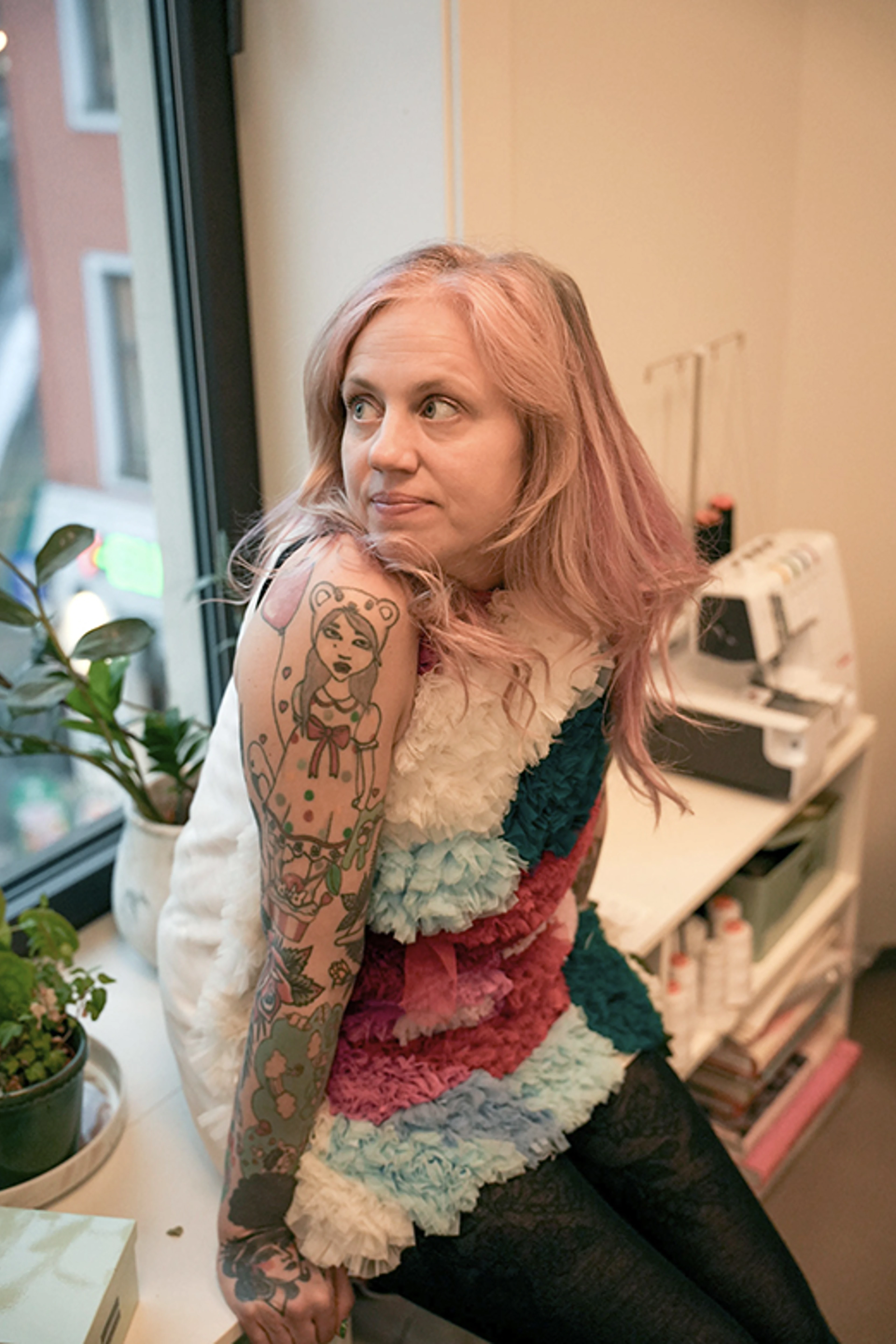 Kvinde med lyserødt hår og tatoverede arme sidder ved et vindue i et arbejdsrum med symaskine og planter.