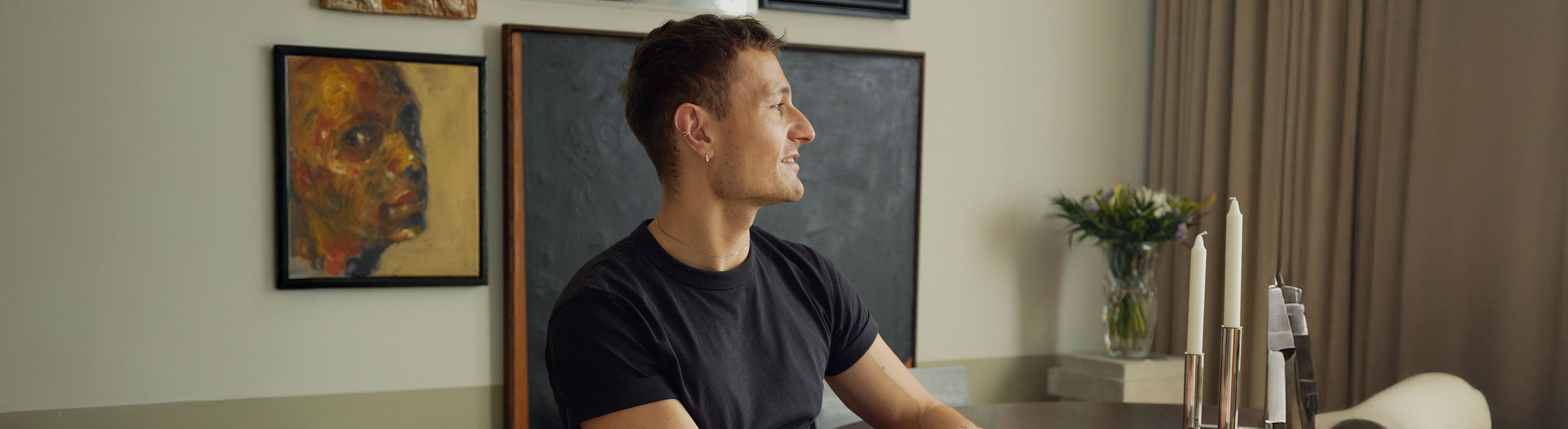 Man in a black T-shirt sits at a dark dining table, holding a mug and looking out the window.