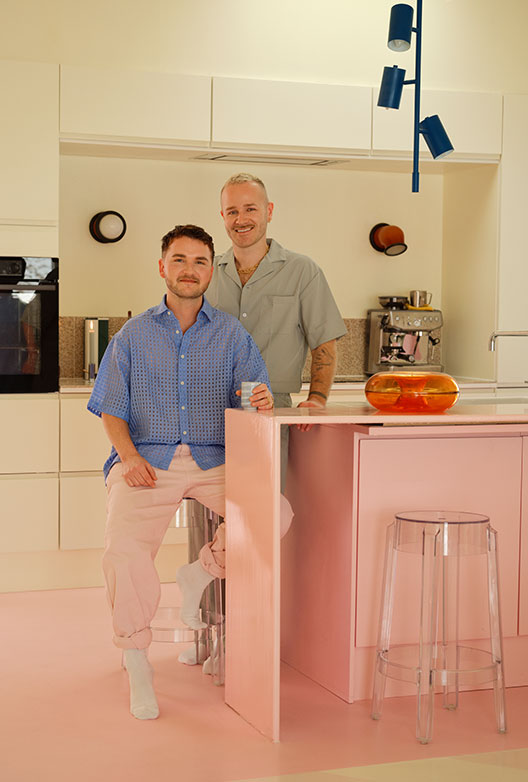 Two men pose in a bright kitchen with a pink floor and kitchen island, surrounded by white cabinets and colorful details.