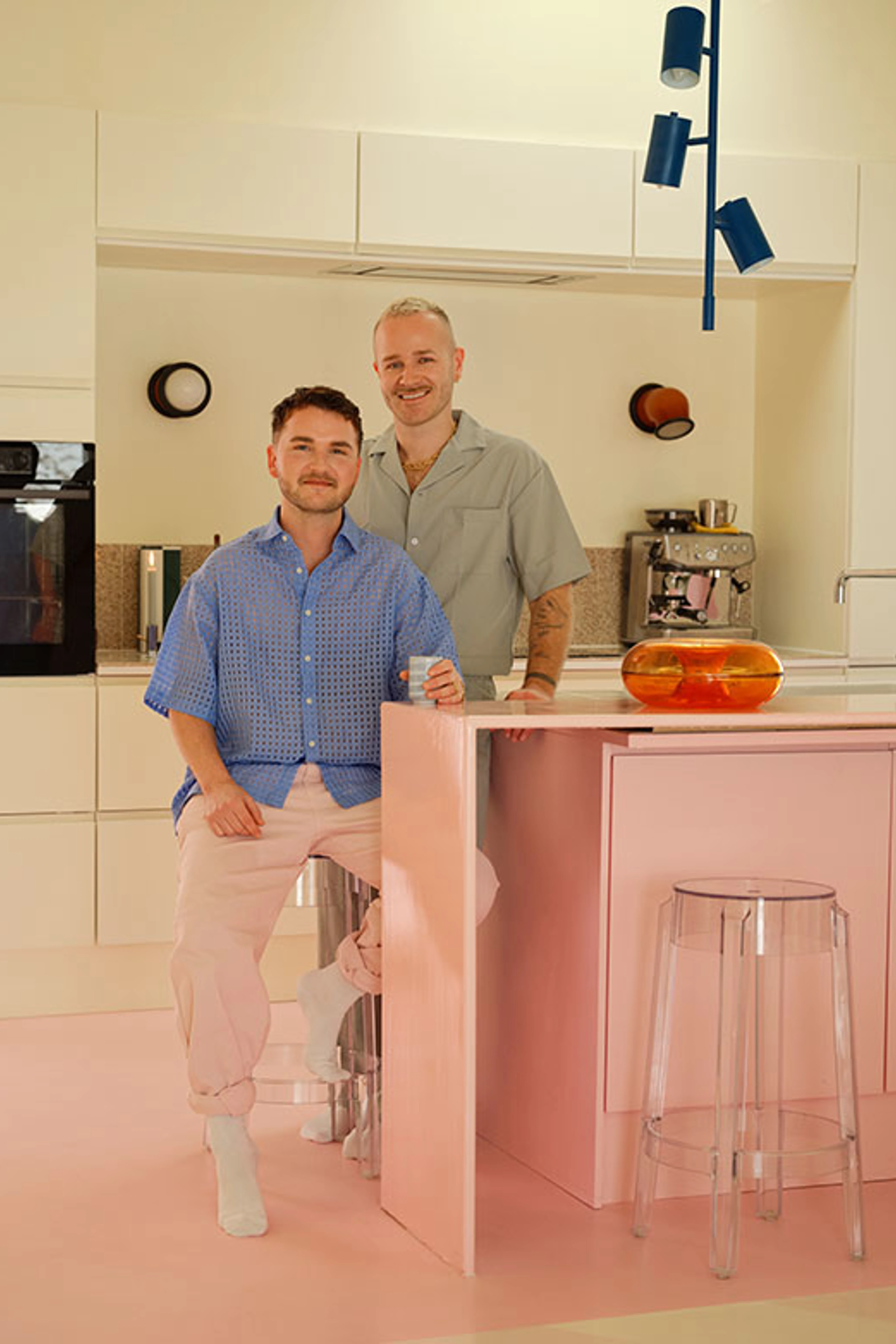Two men pose in a bright kitchen with a pink floor and kitchen island, surrounded by white cabinets and colorful details.