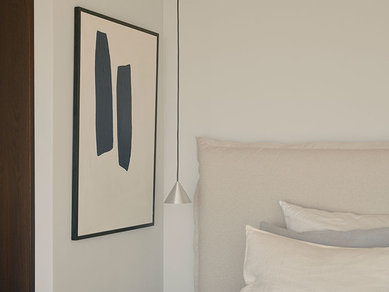 Soveværelse med polstret sengegærde, kunstplakat og pendellampe.