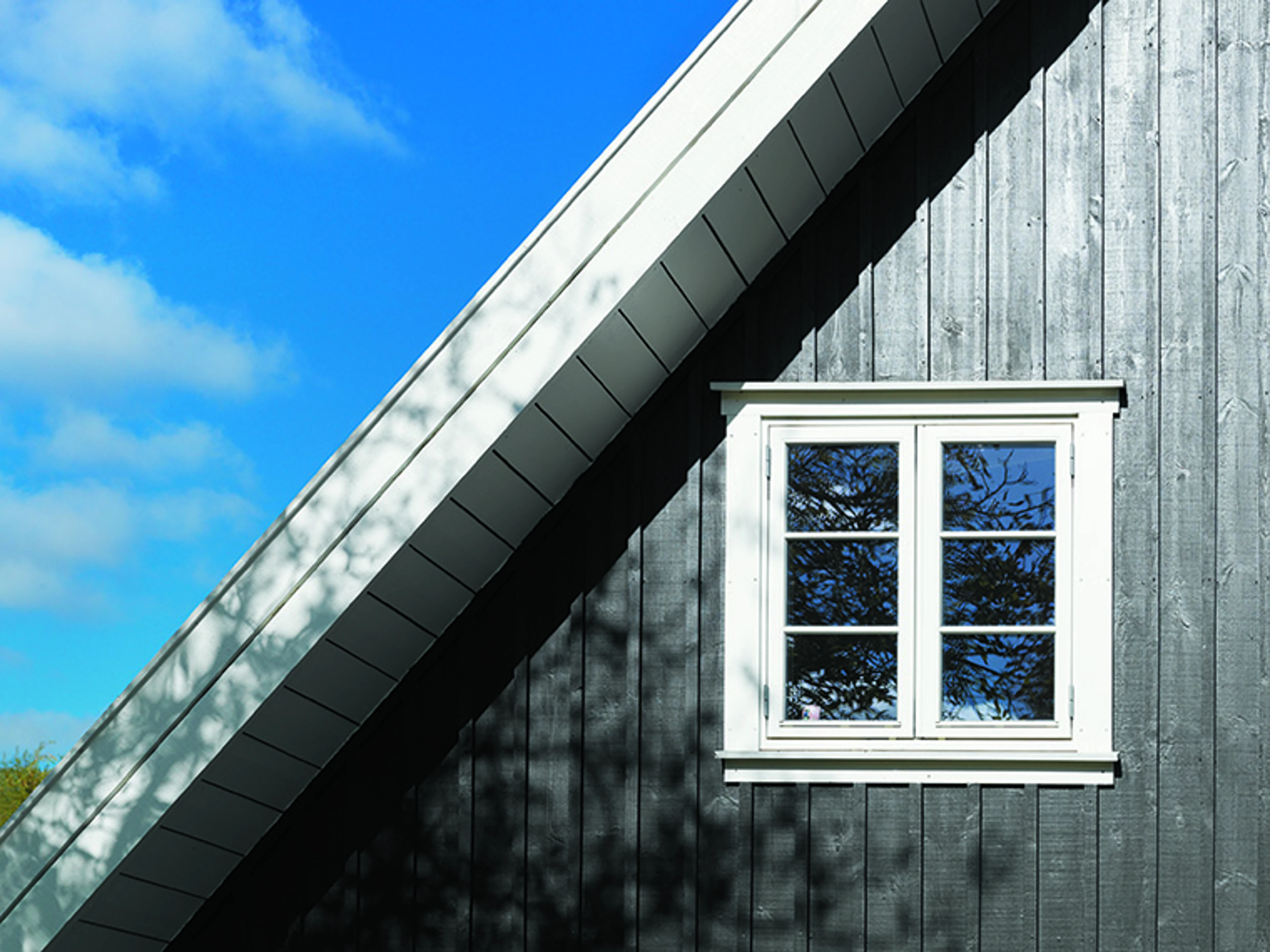 Mørkegrå træfacade med hvidt sprossevindue og hvidt tagudhæng under blå himmel.