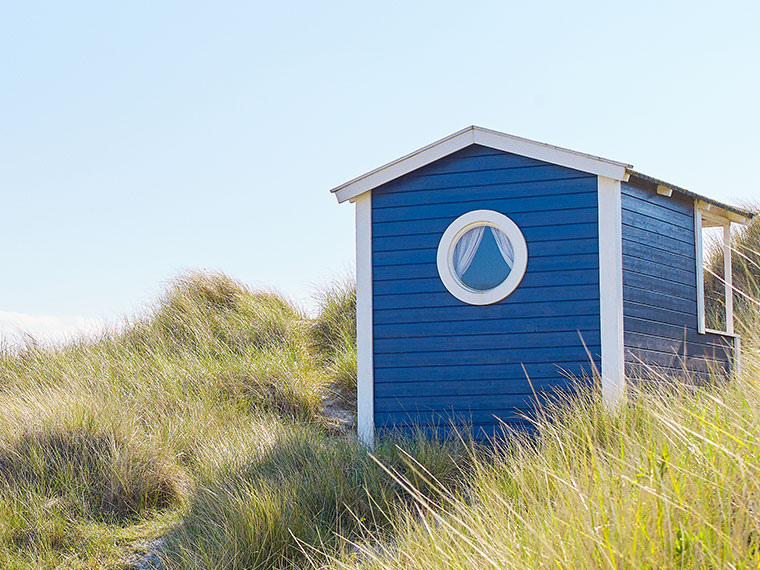 Blå trästuga vid stranden med ett runt fönster, omgiven av dyngräs.