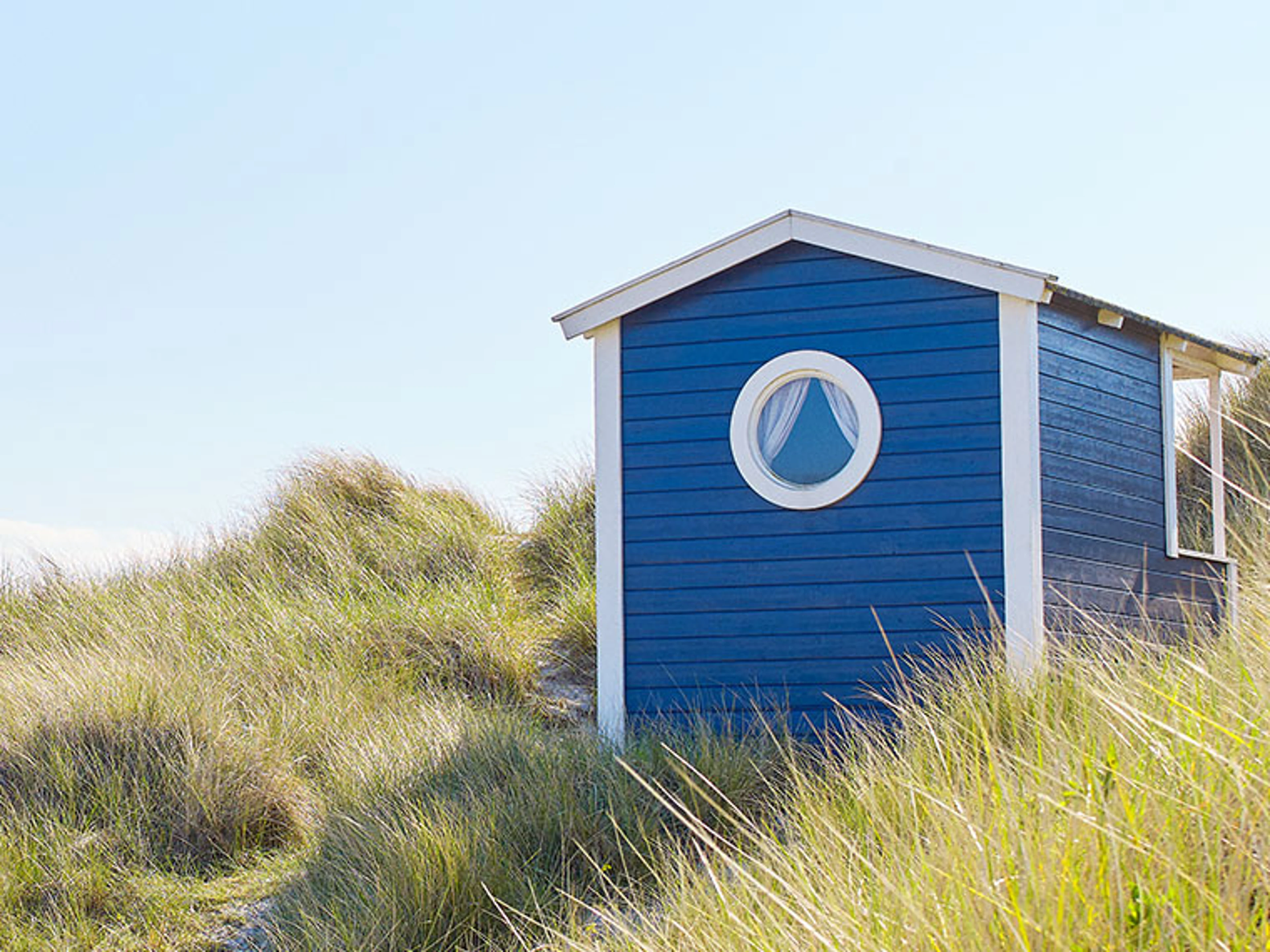 Blå trästuga vid stranden med ett runt fönster, omgiven av dyngräs.
