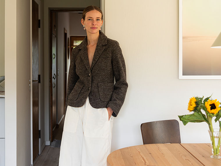 Woman in a tweed blazer and white trousers leans against a white wall by a dining table with sunflowers.