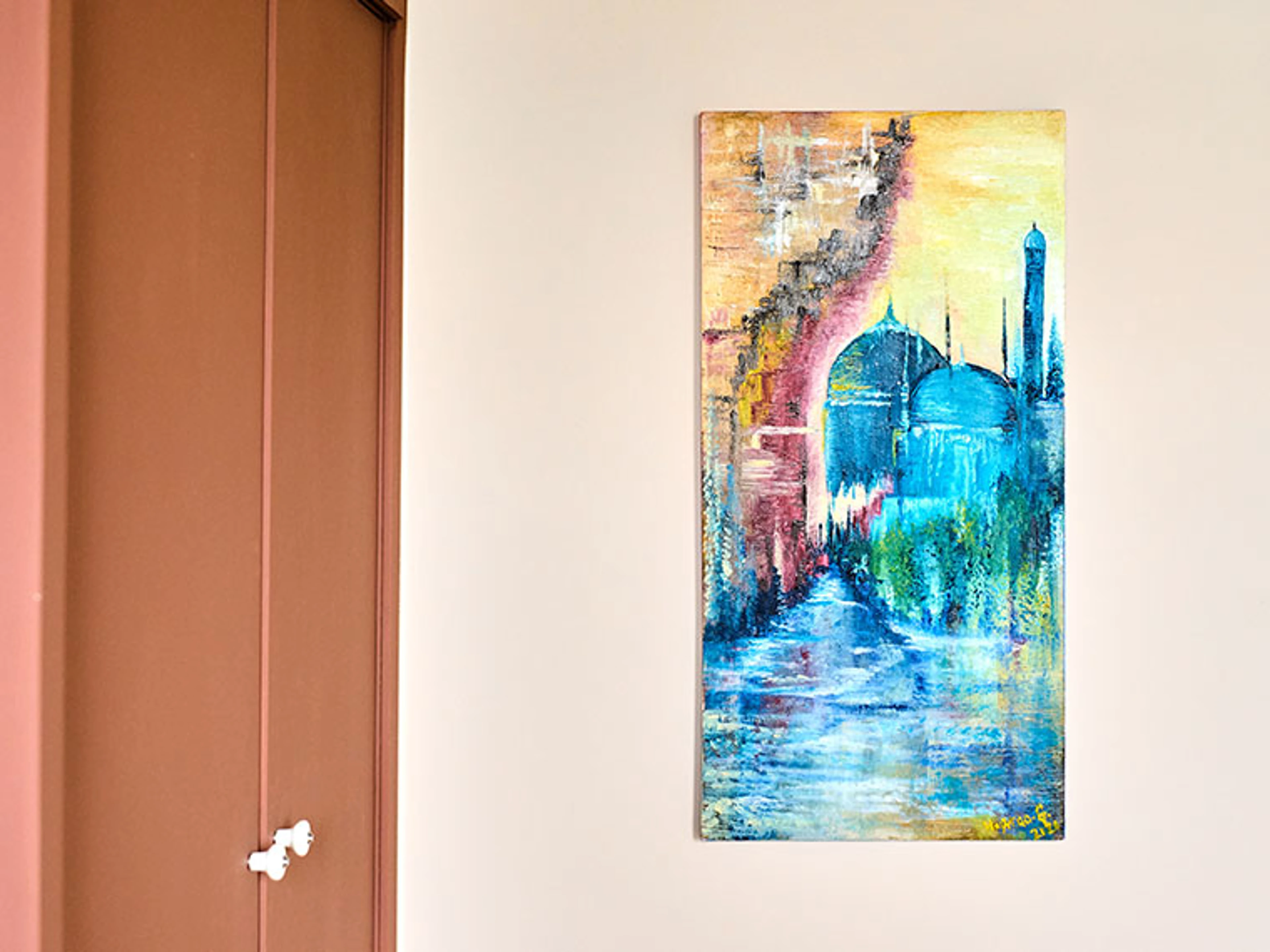 Light-colored wall with a vibrant painting in blue, turquoise, pink, and golden tones beside a terracotta-colored door.