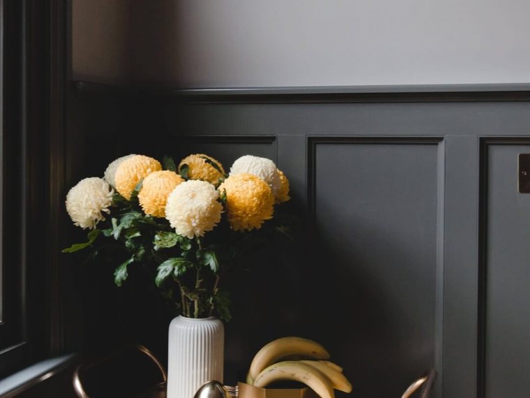 Vase med hvite og lysegule blomster på et bord foran en mørkegrå panelvegg.