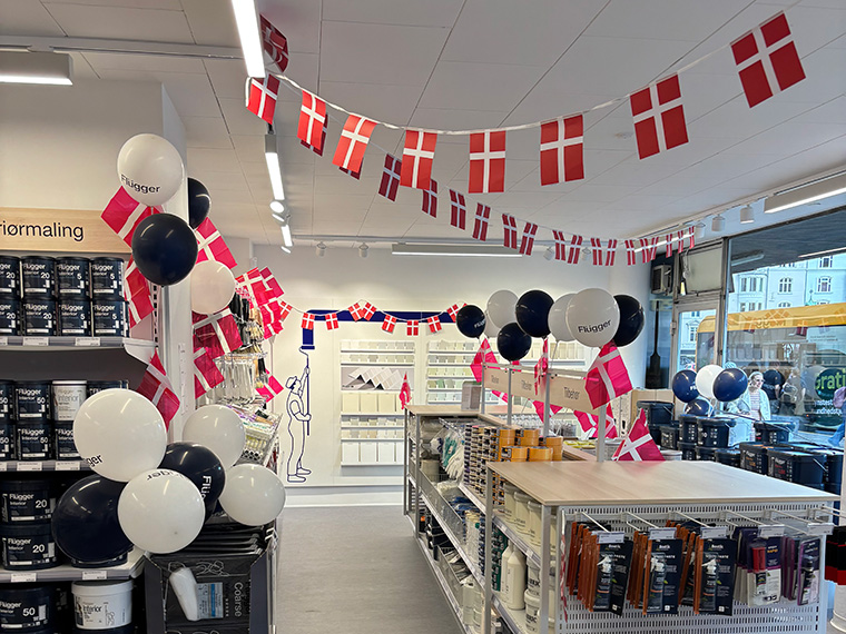 Interiør fra en Flügger-butik dekoreret med danske flag og balloner i rød, hvid og mørkeblå. Butikken indeholder hylder med maling, malertilbehør og udstillingsområder, mens vinduerne ud mod gaden lukker dagslys ind.