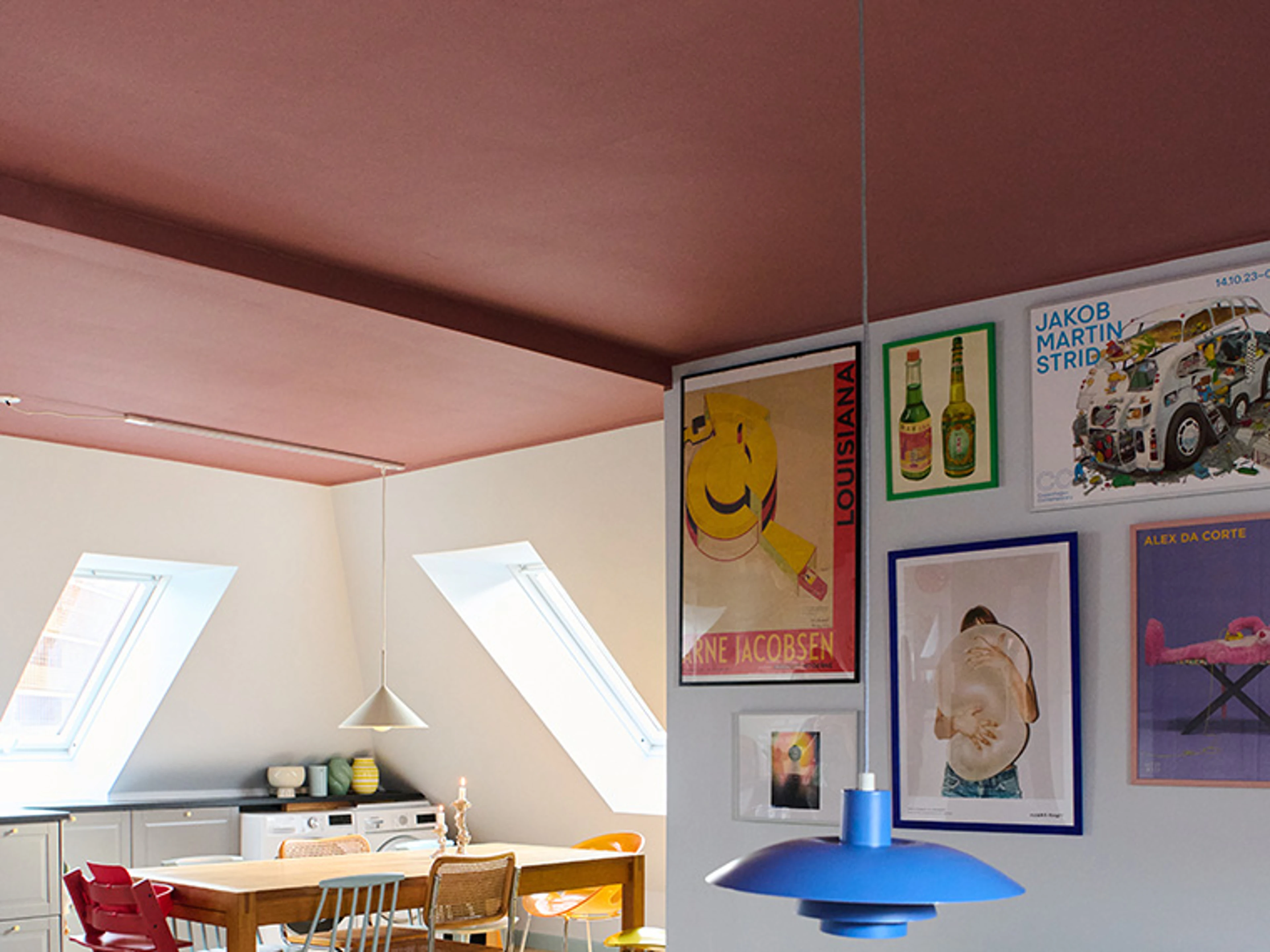 Køkkenalrum med lyse vægge og rosa loft, indrettet med spisebord, stole og væg med farverige plakater.