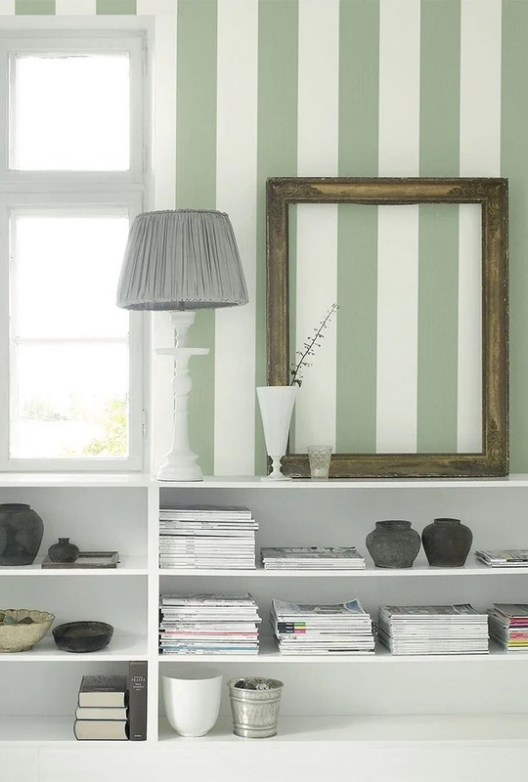 Grønn- og hvitstripet vegg med en hvit reol med magasiner og mørk keramikk, en bordlampe med grå skjerm og en tom forgylt ramme.