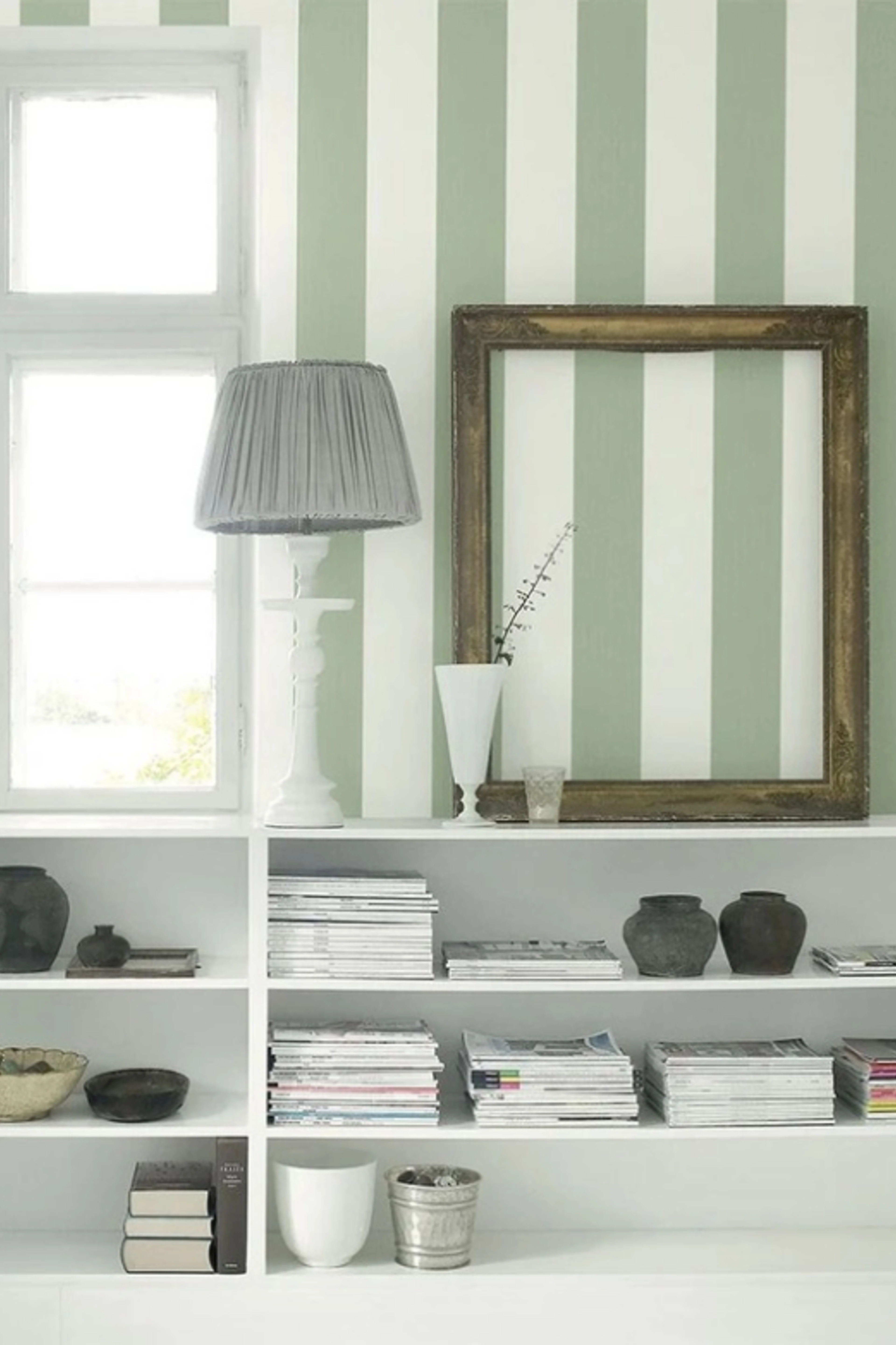 Grønn- og hvitstripet vegg med en hvit reol med magasiner og mørk keramikk, en bordlampe med grå skjerm og en tom forgylt ramme.
