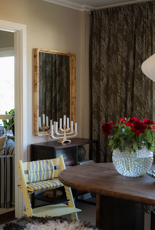 Dining room with beige walls, dark curtains and red flowers on a round wooden table.