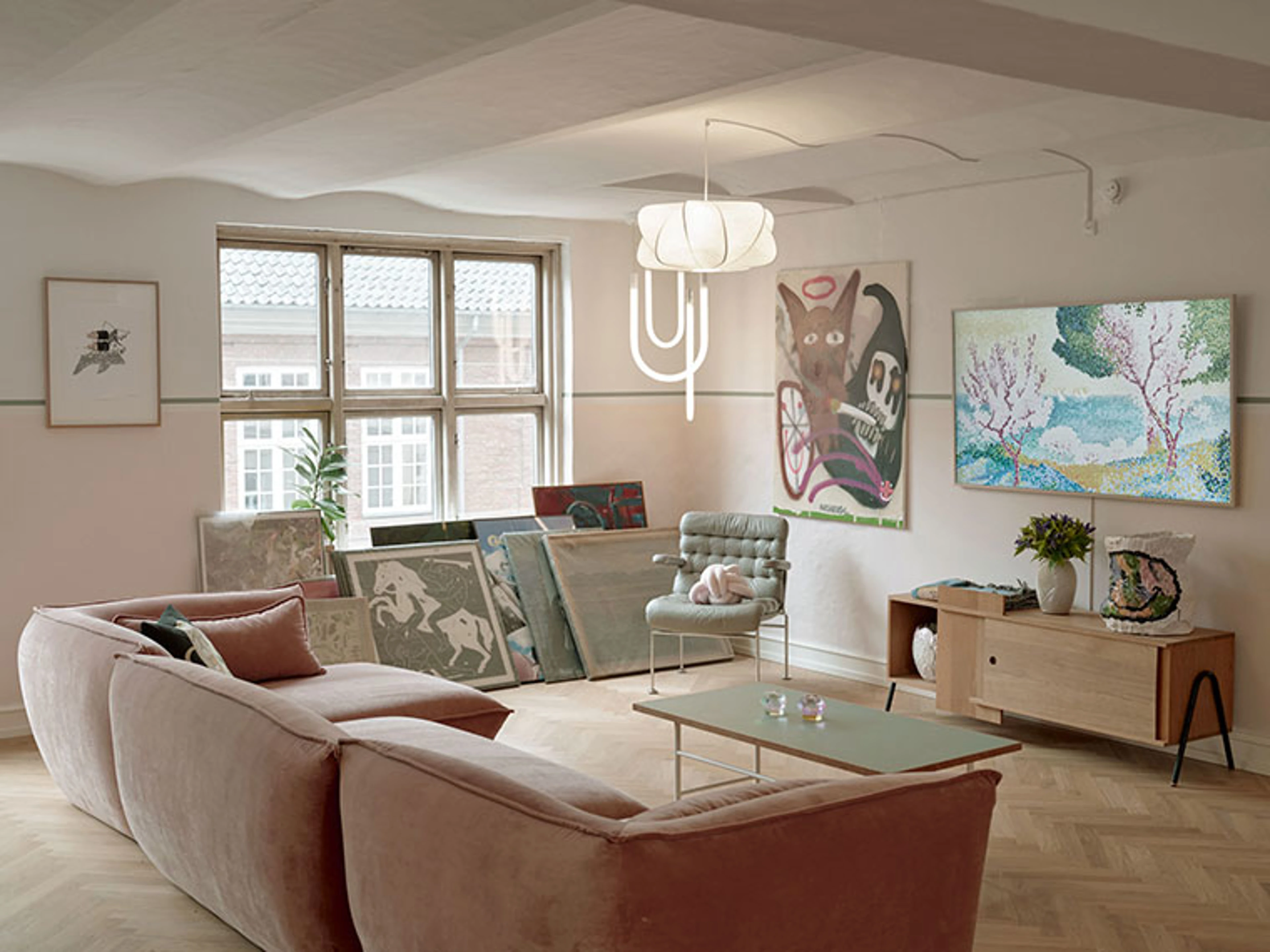 Stue med lyserød sofa, sofabord, kunstværker og loftlampe.