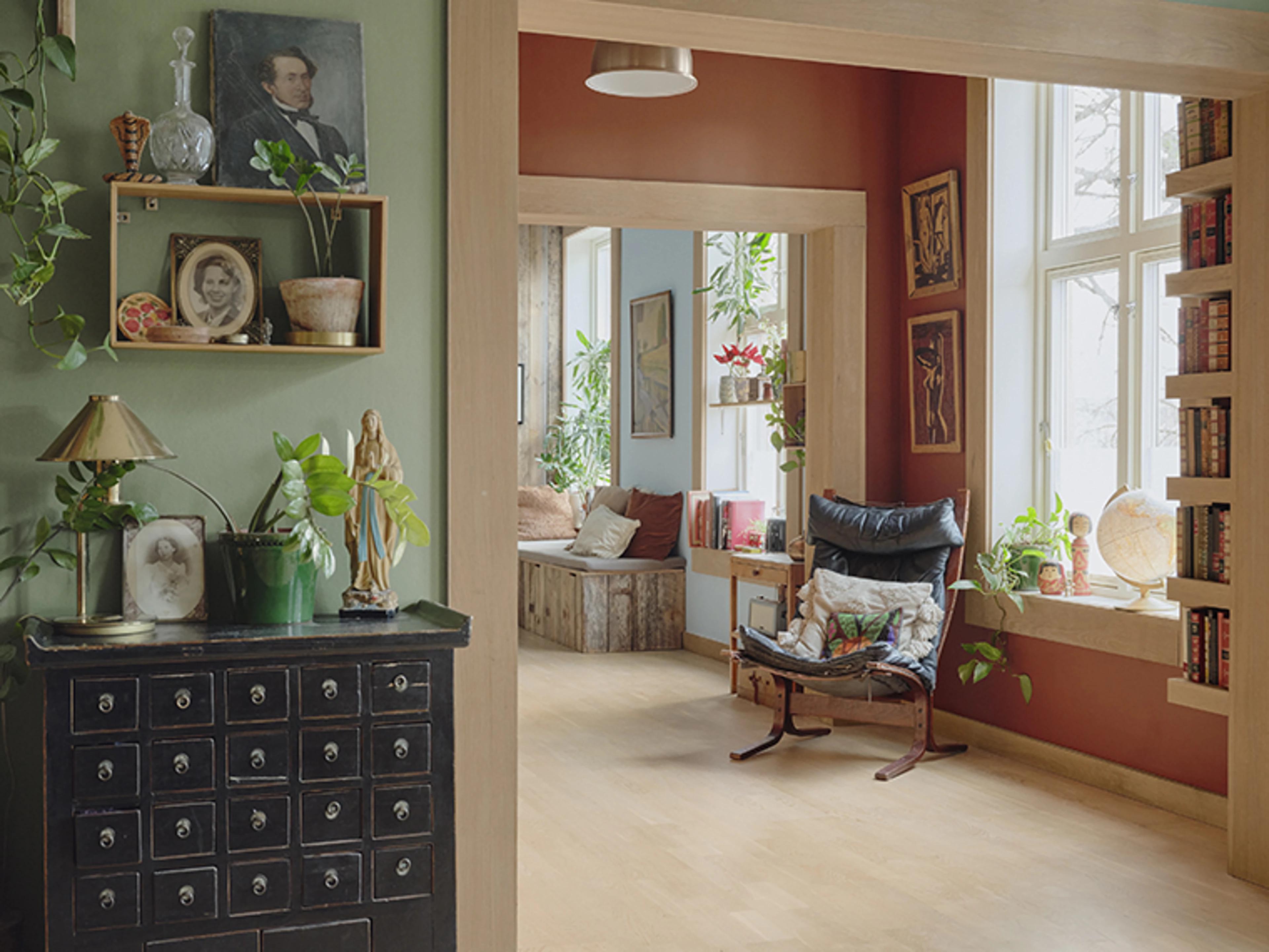 Grøn og rustrød stue forbundet af brede åbninger i lyst træ, indrettet med planter, kunst, bøger og vintage-møbler.