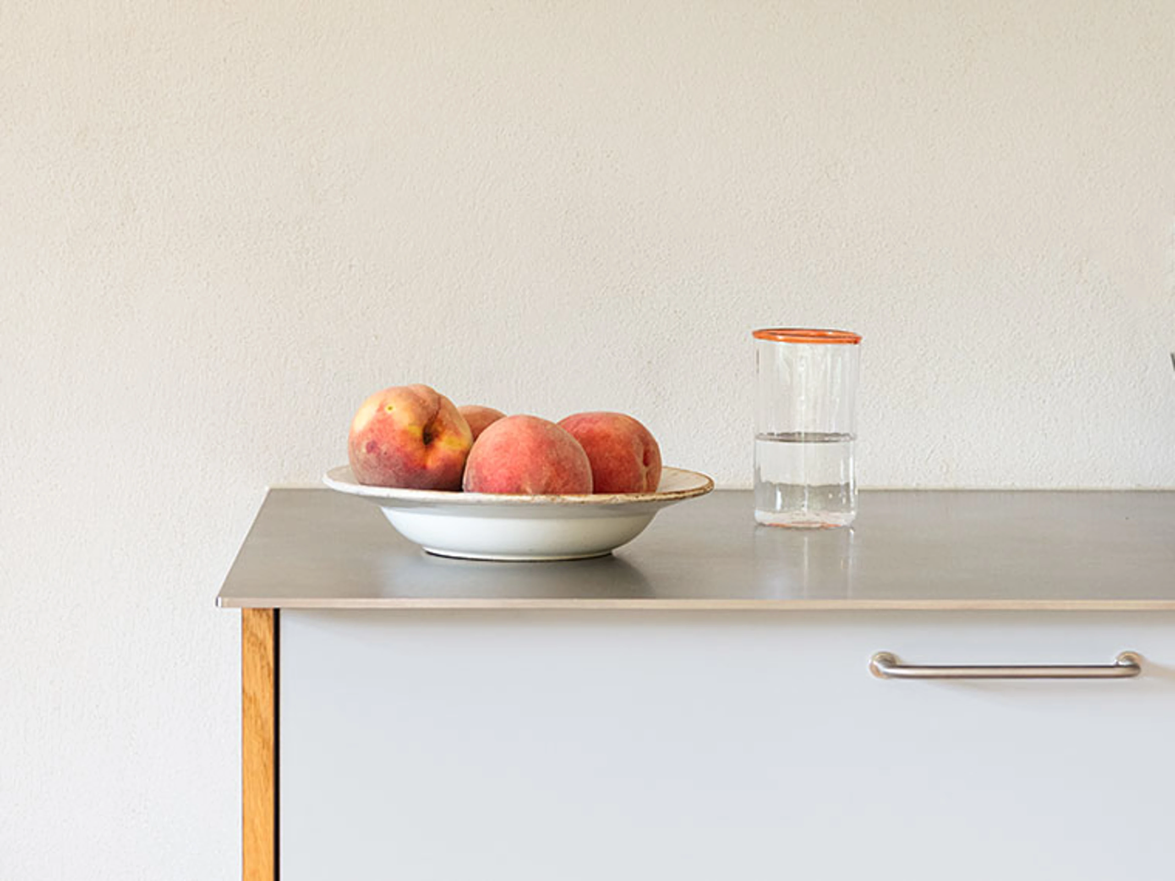 Peaches in a bowl and a glass of water on a steel worktop in front of a white wall.