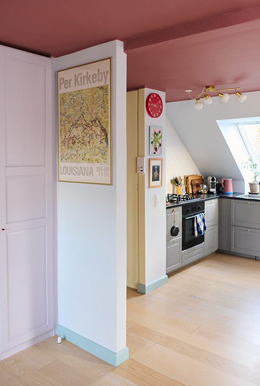 Køkken med lyse vægge og rosa loft, indrettet med skabe, kunst og farverige detaljer i varme nuancer.
