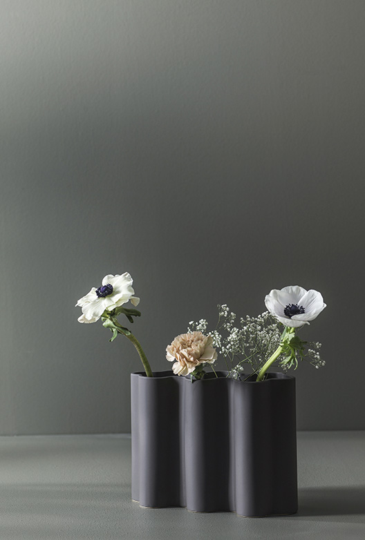 Sorte, cylindriske vaser med hvide og lyse blomster står på en overflade foran en mørk grågrøn væg i et enkelt og stilrent stilleben.