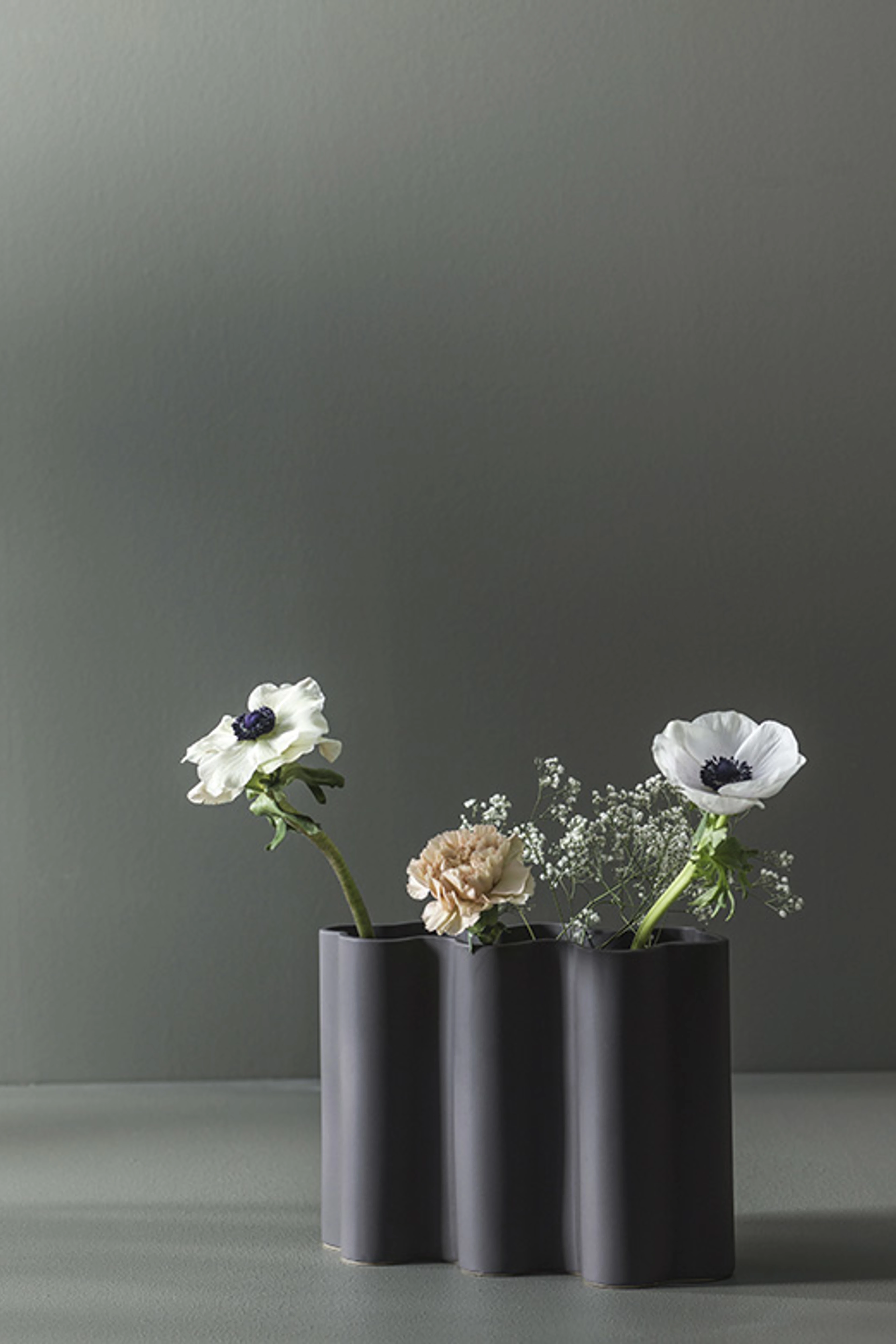 Sorte, cylindriske vaser med hvide og lyse blomster står på en overflade foran en mørk grågrøn væg i et enkelt og stilrent stilleben.