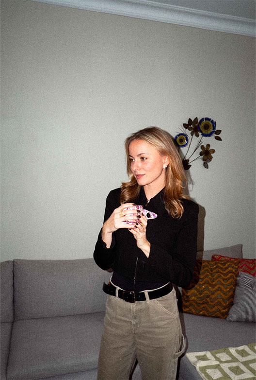 Frederikke står foran en sofa og holder en kop, omgivet af dæmpede farver og tekstiler.