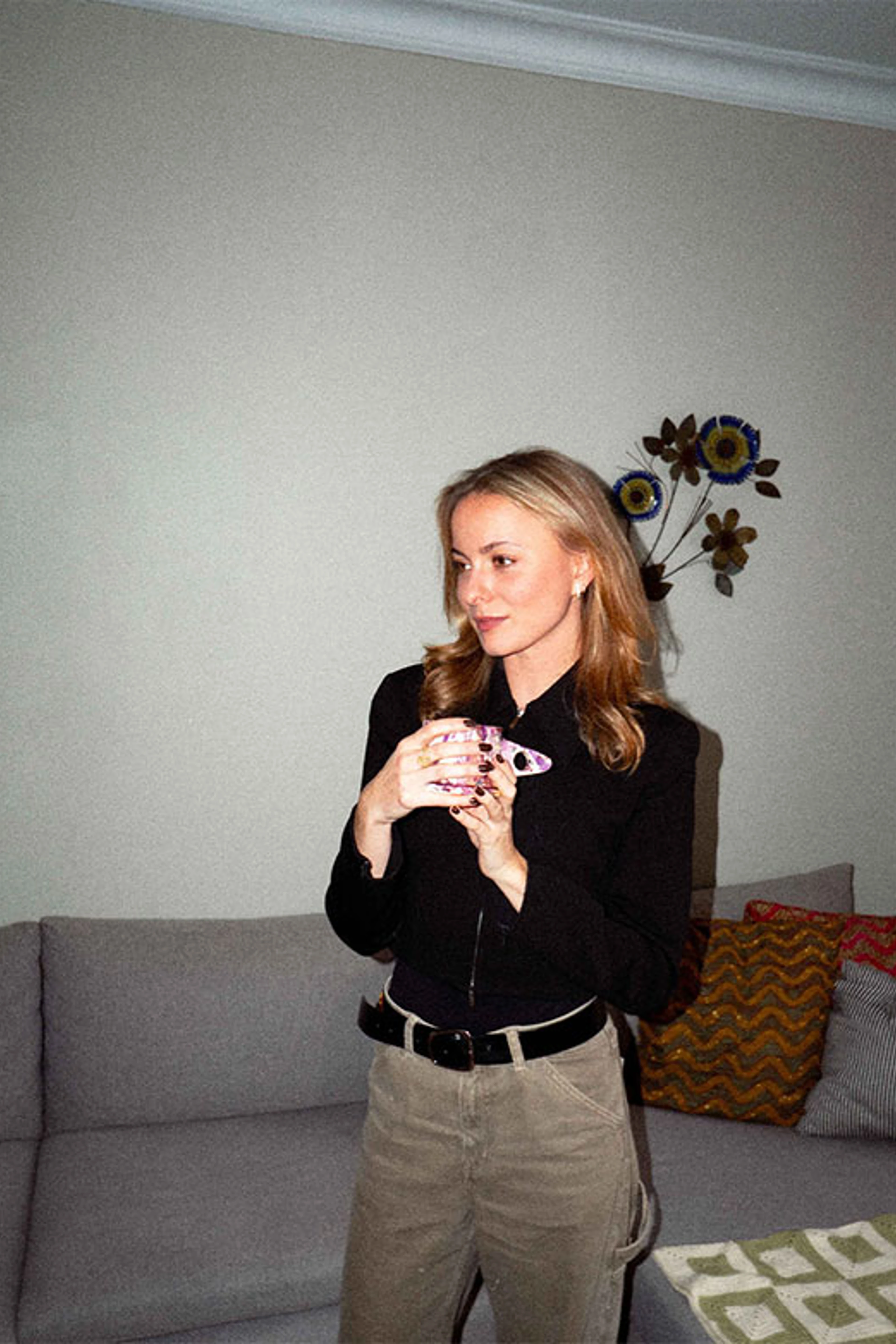 Frederikke står foran en sofa og holder en kop, omgivet af dæmpede farver og tekstiler.