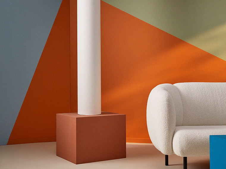 Moderne stuehjørne med en hvid, bouclépolstret sofa foran vægge med geometriske farvefelter i orange, blå og lysegrønne nuancer. En hvid søjle står på en terrakottafarvet sokkel og understreger det grafiske designudtryk.