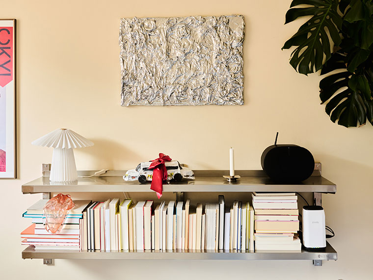 Væghængt stålreol med bøger, lampe, højttaler og dekorative genstande.